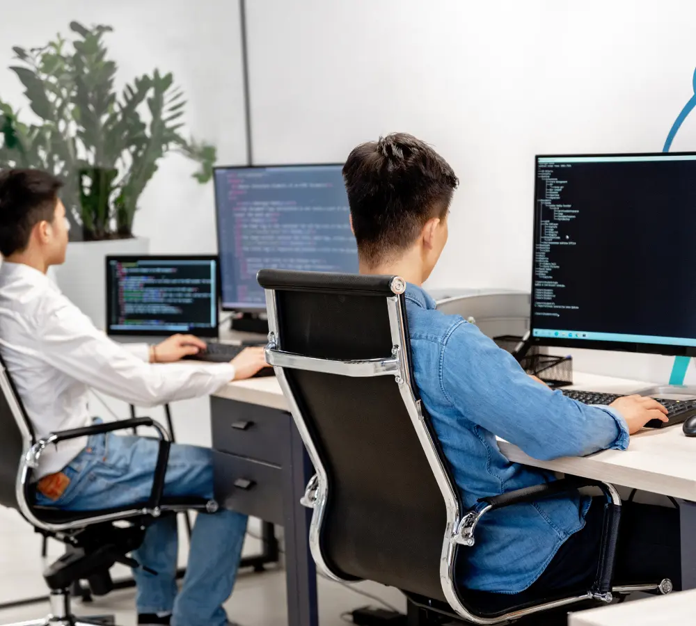Valencia interior 2 A programmer sitting at an office desk, with their back to the camera, focused on a large monitor displaying technical code, representing the development and continuous implementation of AI assistant solutions for IT support.